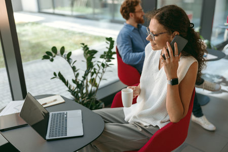 Woman freelancer talking on phone with client while working on laptop in modern coworkingの写真素材