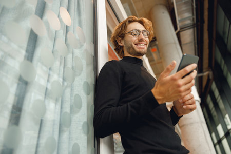 Handsome male freelancer holding phone while standing in office during break timeの写真素材