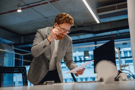Focused entrepreneur working with documents standing near desk in modern officeの写真素材