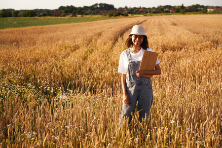 Smiling female farmer standing with clipboard on wheat field during harvesting. Agricultural conceptの写真素材