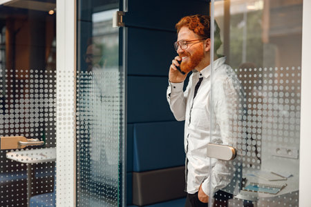 Bearded male freelancer in eyeglasses is talking phone standing in modern coworking spaceの写真素材
