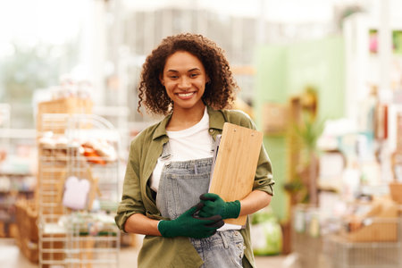 Smiling woman gardener standing with clipboard on garden center background and looks cameraの写真素材