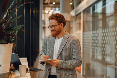 Smiling male manager working on laptop and making notes while standing on office backgroundの写真素材