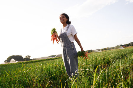 Smiling female farmer holding freshly picked carrots standing in field. Agro industry conceptの写真素材