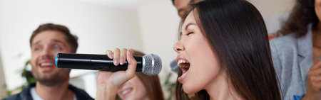 Home party. Young beautiful asian girl holding microphone and singing while playing karaoke with her best friends at home. Group of happy people having fun together. Selective focusの写真素材