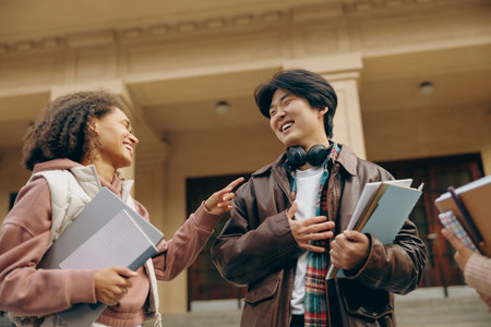 Young students talking and laughing together outdoor standing near campus and holding books