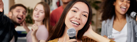 Bring joy. Young pretty asian woman holding microphone and singing while having fun with friends at...