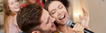 Sing together. Young excited couple or friends holding microphone and singing together while playing karaoke with friends at home. Group of people having fun at karaoke home partyの写真素材