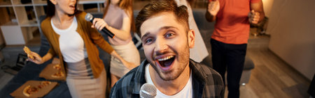 Show your singing skills. Happy young guy singing with microphone while playing karaoke with friends at home, dancing in the modern apartmentの写真素材
