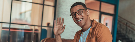 Smiling freelancer working on laptop while sitting in coworking background and waving hi on cameraの写真素材