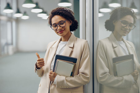 Pretty female manager in eyeglasses standing on office background and showing thumb upの写真素材