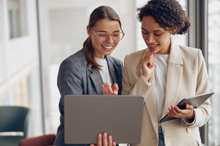 Stylish female managers standing in modern office while working on laptop and digital tabletの写真素材