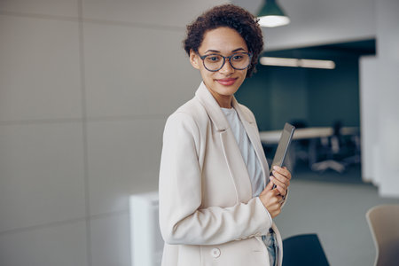 Smiling female manager with digital tablet standing on modern office background and looks cameraの写真素材
