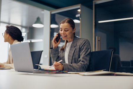 Smiling business woman working on laptop while sitting in office and talking by phoneの写真素材