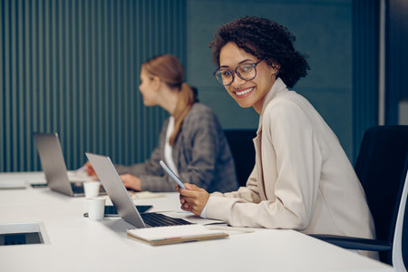 Smiling female freelancer working on laptop and holding phone while sitting in modern coworkingの写真素材
