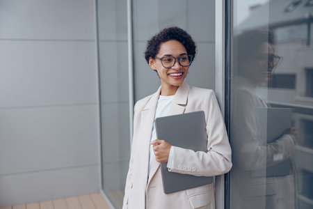 Smiling businesswoman holding laptop while standing on office terrace and looks awayの写真素材