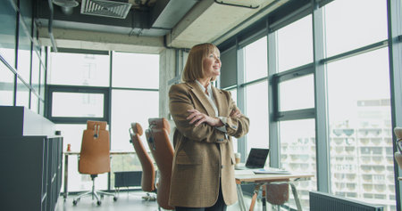 A confident mature businesswoman standing with arms crossed in a bright modern office, showcasing leadership and success in a professional corporate environmentの写真素材