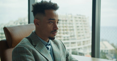 A focused businessman in a tailored suit sitting in a modern office with large windows and an urban cityscape in the background, showcasing professionalism and determination.の写真素材