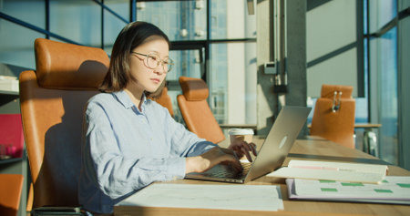A focused young professional woman working on a laptop in a modern office with sunlight streaming through large windows, surrounded by office furniture and documentsの写真素材