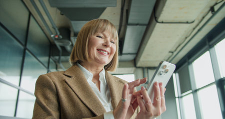 A mature businesswoman using a smartphone in a modern office environment, focusing on professional tasks or communication, highlighting confidence and technological proficiencyの写真素材