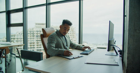 A professional businessman working on a computer and taking notes in a modern office with large windows, showcasing focus and productivity.の写真素材