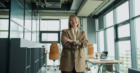 A confident mature businesswoman standing with arms crossed in a contemporary office setting, emphasizing professionalism and leadership in a modern corporate workspaceの写真素材