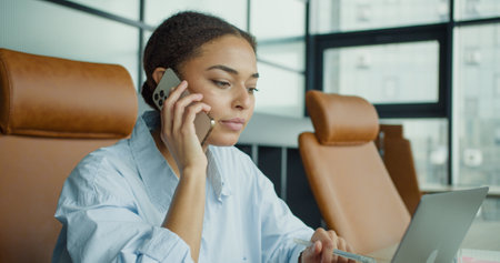 A young woman multitasking in a modern office, talking on the phone while holding a pen, looking focused and engaged at her workspace with a laptopの写真素材