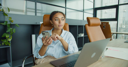 young woman checking her smartphone while working on a laptop in a modern office, sitting at a desk with notebooks, a glass of water, and stylish leather chairsの写真素材