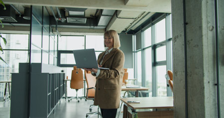 A mature businesswoman holding a laptop in a modern office environment, showcasing mobility and...