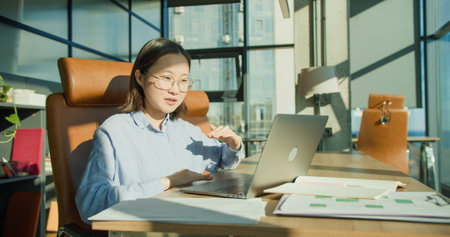 A woman in glasses having a video call in a modern office with a laptop and coffee, gesturing expressively and smiling under natural sunlightの写真素材