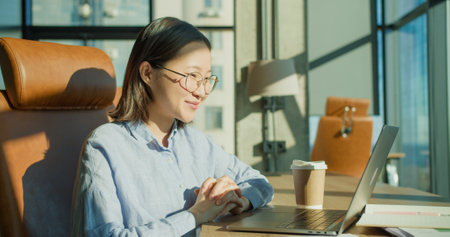 A young woman relaxing with her hands behind her head in a modern office with a laptop and coffee cup on the desk, enjoying sunlight through large windowsの写真素材