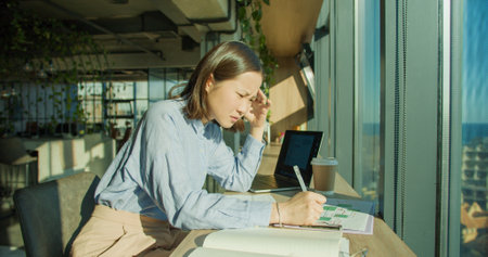 A focused young woman in a modern office environment analyzing documents and working on a laptop, surrounded by bright sunlight and an urban cityscapeの写真素材