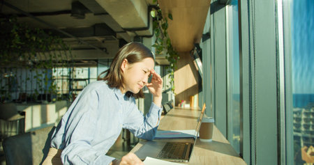A thoughtful young woman in a modern office struggles with fatigue as sunlight streams through large windows, surrounded by a laptop, coffee, and notepadの写真素材