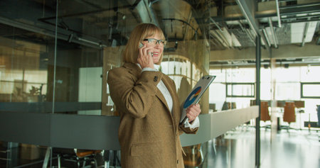 A professional businesswoman in a modern office environment, confidently discussing on the phone while holding a clipboard with data charts, standing near a glass partitionの写真素材