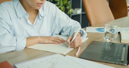 A close-up of a focused young woman writing notes at a desk in a modern office, with glasses, a pen,...