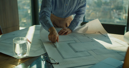 A detailed architectural plan being reviewed on a wooden table by a professional in a modern office,...