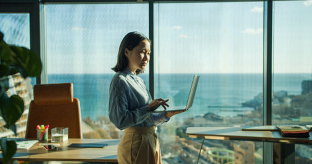 Young professional woman using a laptop in a bright office with a panoramic ocean view, working while standing near large windows in a modern workspaceの写真素材