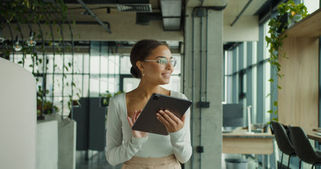 Happy young woman in stylish outfit using a digital tablet in a modern office with plants and natural light through large glass windowsの写真素材