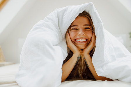 A woman lays in bed under a white blanket, smiling and feeling comfortableの写真素材