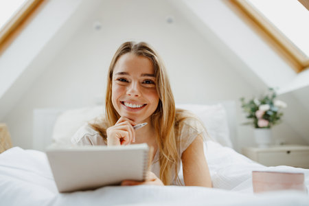 Blond happy woman making notes in note pad while sitting on bed at homeの写真素材