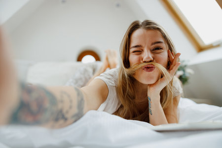 A woman is sitting comfortably on the bed, smiling and taking a selfie with her phoneの写真素材