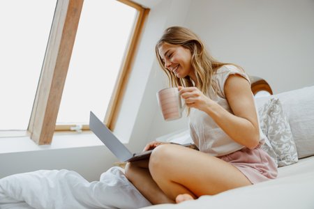 A woman is sitting comfortably on a bed with her legs crossed, using a laptop and sipping coffeeの写真素材