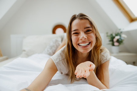 A happy woman is laying on a bed, blowing a kiss with her hand while her arm and finger are extendedの写真素材