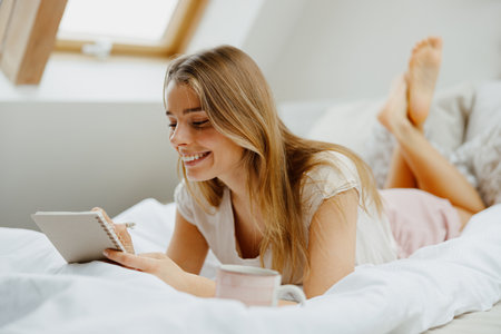 A woman in pajamas is seated on a wooden bed and making notes in note padの写真素材