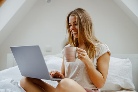Woman is sitting on a bed using a laptop computer while holding a cup of coffeeの写真素材