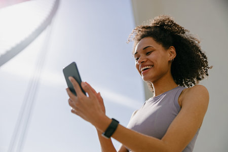 Smiling woman athlete in sportswear standing outdoors after training and use mobile phoneの写真素材