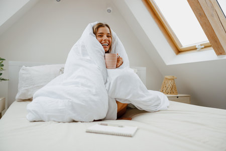 Happy woman is sitting on a bed wrapped in a white blanketの写真素材