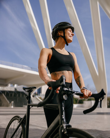 Young woman cyclist in protective gear standing with her bike while training outdoors and looks awayの写真素材