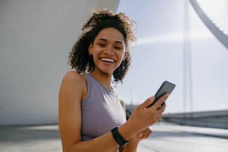 Young smiling woman in sportswear is resting after a workout outside while standing with smartphoneの写真素材
