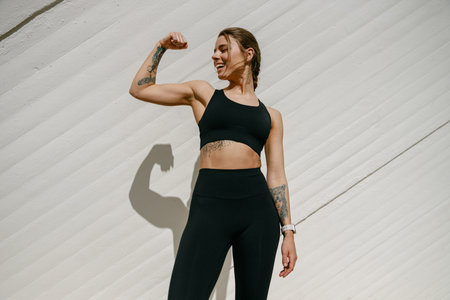 Smiling female model in sportswear showing biceps standing outdoors and looks away